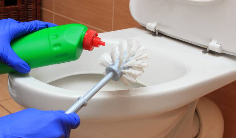 Hand in blue gloves holding a green bottle with red cap and a brush in front of toilet