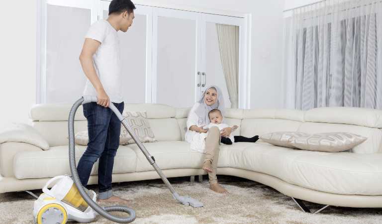 Man in white t-shirt and black pant holding a vacuum cleaner and a woman in sitting on her sofa with her kid.